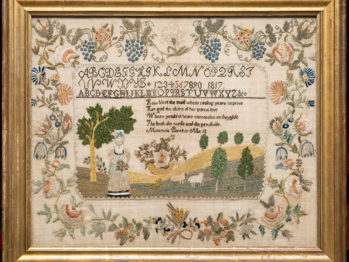 Needlework of a country scene surrounded by flower imagery in a gold frame. 